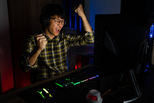 Захоплений азіатський чоловік-програвач сидить за столом, грає і виграє в онлайн відеоіграх на комп'ютері і смартфоні, концепції технологічних ігор кіберспорт або чемпіонату
. - Фото, зображення