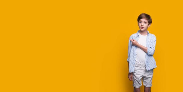 Fiatal fehér fiú pólóban pózol meglepve egy sárga stúdió falán, szabad hellyel, mutogatva valamire mutatóujjal. - Fotó, kép