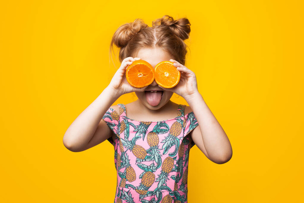 Kaukasisches kleines Mädchen mit blonden Haaren und Sommerkleid posiert mit aufgeschnittenen Orangen auf den Augen und geöffnetem Mund auf einer gelben Studiowand - Foto, Bild