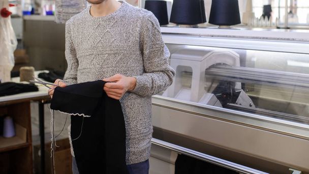 コーンの黒い糸で編んだ機械の近くで編んだ布を分析している若い男が  - 写真・画像