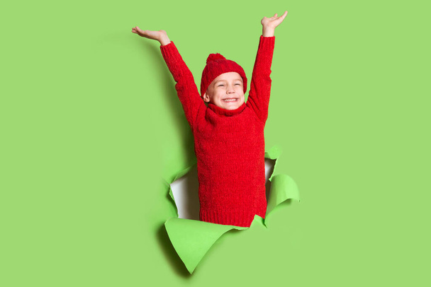 cheerful kid boy in a red sweater poses through torn paper hole  wall. Effect of torn paper. merry Christmas - Foto, imagen
