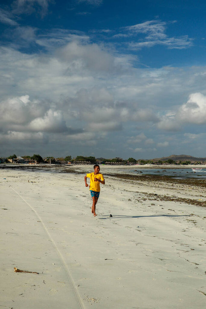 Бегущий человек бегает по пляжу. Индонезийский подросток в шортах и желтая футболка, бегущая вдоль пляжа. Юный спортсмен тренируется у моря. Подходящая модель фитнеса для юношей летом. - Фото, изображение