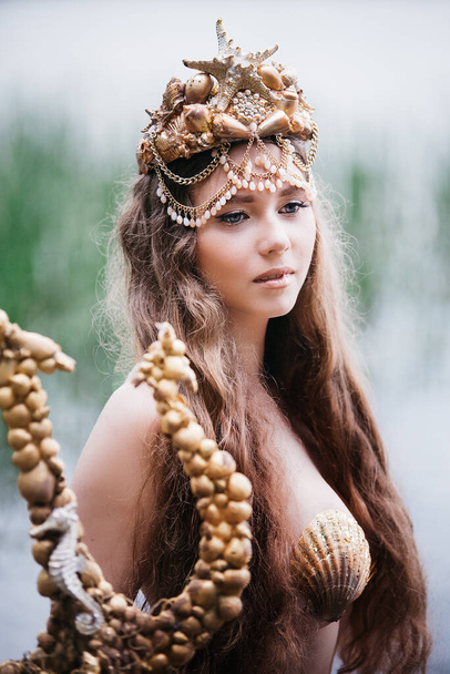 Fantasy woman real mermaid with trident myth goddess of sea with golden tail sitting in sunset on rocks.. Gold hair crown shells pearls jewelry. Mermaid sitting on shore. fantasy concept. - Photo, image