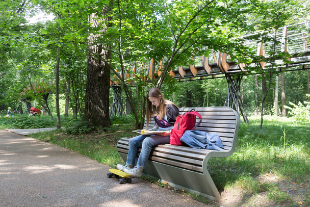 Ένα έφηβο κορίτσι διαβάζει ένα βιβλίο σε ένα παγκάκι σε ένα πάρκο της πόλης. - Φωτογραφία, εικόνα