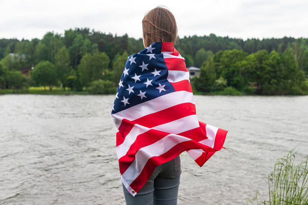 Young woman holding American flag on lake background. United States celebrate 4th of July, sad lonely girl close up - 写真・画像