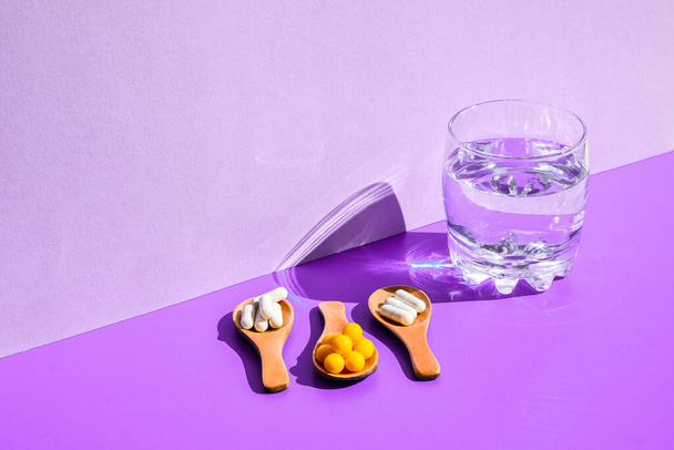 Glass of water and pills on wooden spoon on purple background with hard light and shadows. Modern isometric creative minimalism concept. Vitamins and prebiotics, probiotics. Autumn vitamin dose. - Photo, Image