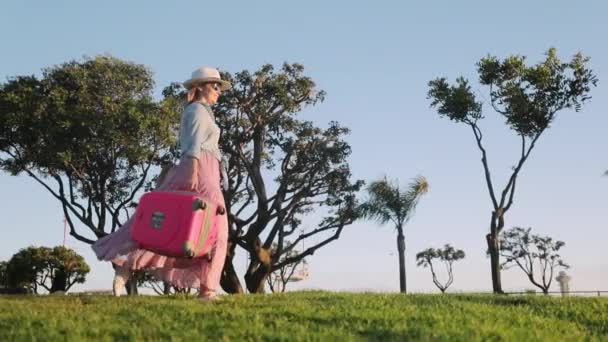 陽気な女性は彼女の旅行に興奮し、日没時にピンクの旅行バッグとジャンプします - 映像、動画