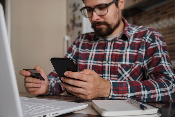Handsome man using credit card to pay online with smartphone. Shopping Online, Banking system. guy in a plaid shirt sitting in his home kitchen - Foto, immagini