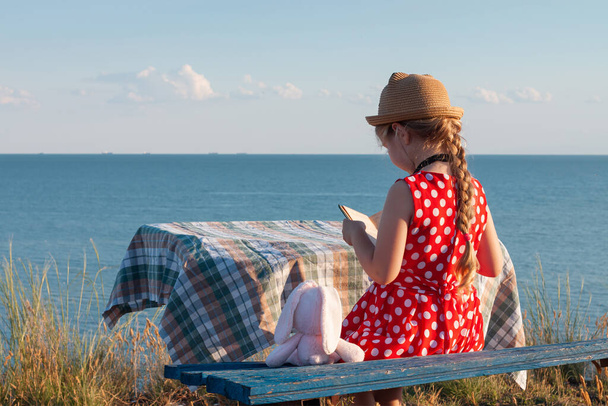 Child girl in hat and polka-dot dress sitting on vintage bench reading a book. Kid looking at Holy Bible in hands and praying on sea lanscape background. Friendship peace religion faith hope concept. - 写真・画像