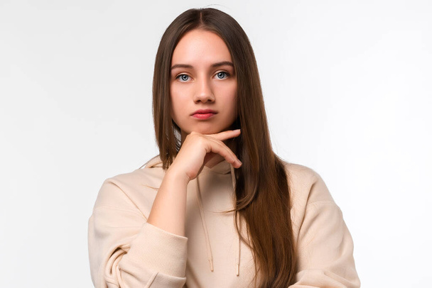 Photo of Pensive young woman with long chestnut hair standing with hand raised on chin and looking thoughtfully into a camera isolated over white background. Concept of thinking - Foto, Imagen