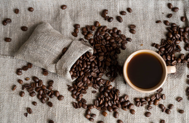 Coffee scattered from a linen bag and a cup of black coffee Espresso or Americano standing on a wooden table. Fresh arabica coffee beans on linen textile. Aromatic freshly brewed coffee. - Photo, image