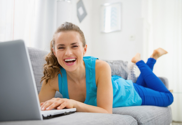 Усміхнена молода жінка лежить на дивані і використовує ноутбук
 - Фото, зображення