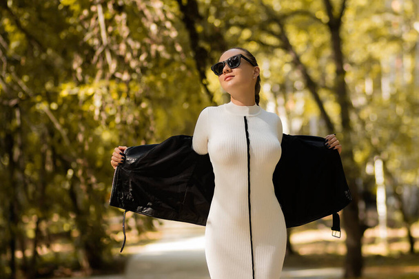 Femme mince en robe blanche tendance et lunettes de soleil décollant veste en cuir le jour ensoleillé d'automne dans le parc - Photo, image