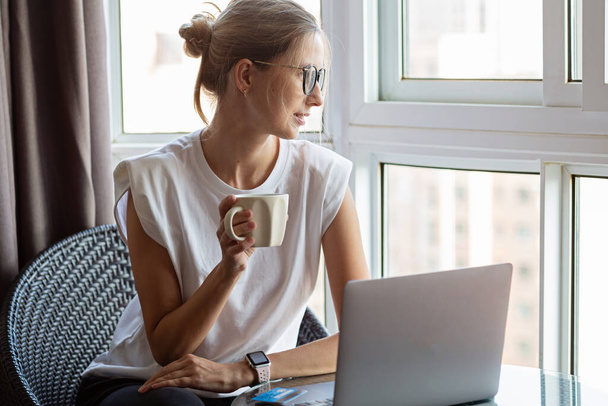 Young woman wearing smartwatch, drinking hot coffee and using laptop. Female working on computer pc at home during covid-19 coronavirus quarantine. Stay home, social distance concept. High quality photo - Foto, Imagen