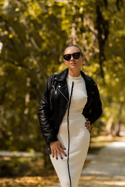 Slim female in trendy white dress and sunglasses taking off leather jacket on sunny autumn day in park - Foto, imagen