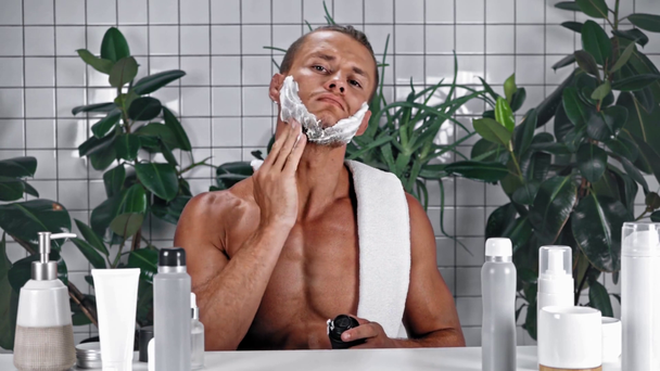 Άνδρας με πετσέτα που συμπιέζει αφρό ξυρίσματος και εφαρμόζεται στο πρόσωπο στο μπάνιο - Πλάνα, βίντεο