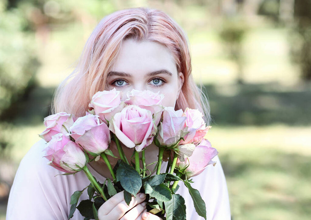 Beautiful girl with blue eyes sniffs pink roses, outdoors. Summertime. - Photo, Image