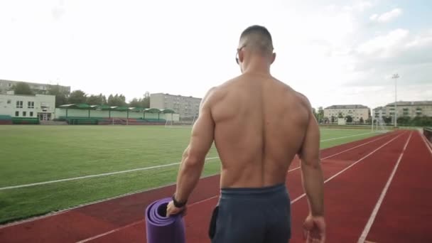 Een bodybuilder in sportbroek met een mat in zijn hand gaat naar het veld van het stadion voor training. Zicht van achteren. Een close-up. De camera volgt hem. - Video