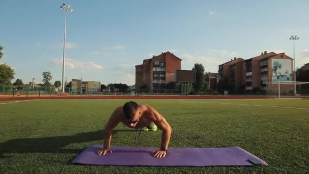 Muscular man with a naked torso in sunglasses doing push-ups on a sports mat at the city stadium. Close-up. Front view. The camera follows the movements. Sportsman uses mobile app on his phone for - Footage, Video