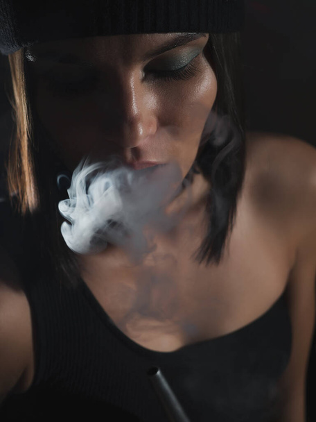 Attractive woman in hat exhaling hookah smoke while resting in dark room at home - Fotó, kép