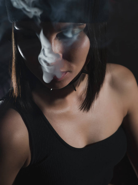 Aantrekkelijke vrouw in hoed uitademen hookah rook tijdens het rusten in donkere kamer thuis - Foto, afbeelding