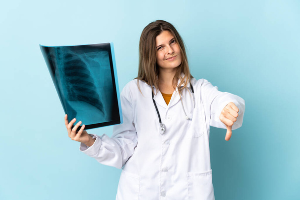 Junge Ärztin hält Röntgenbild über isoliertem Hintergrund und zeigt Daumen nach unten mit negativem Ausdruck - Foto, Bild
