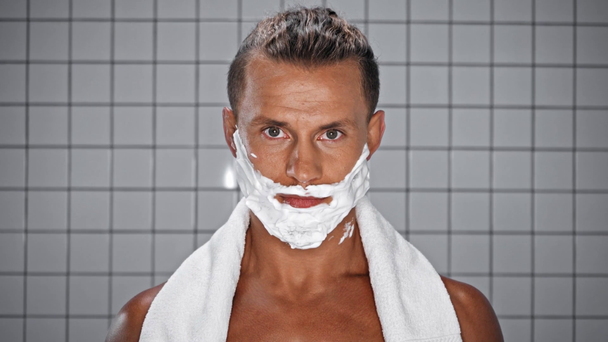 Hombre con espuma de afeitar en la cara mirando a la cámara en el baño - Metraje, vídeo