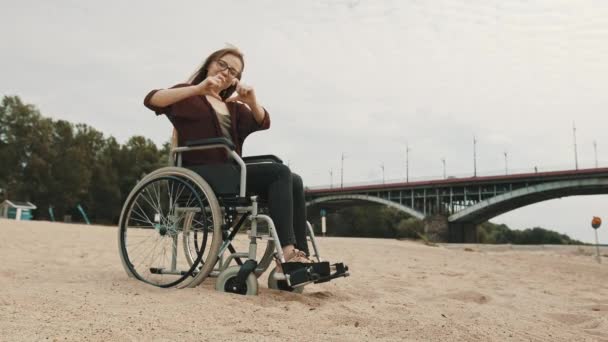Suport für behinderte Menschen. Junge Frau im Rollstuhl an der sandigen Flussküste zeigt Herz mit Händen - Filmmaterial, Video