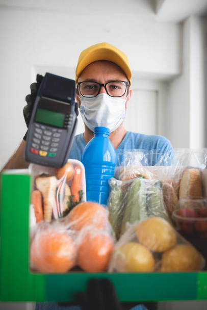 Delivery guy with protective mask holding box / bag with groceries and POS for contactless payment. - Photo, Image