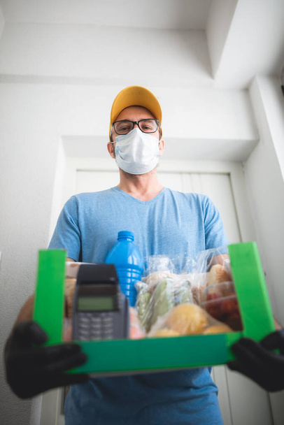 Delivery guy with protective mask holding box / bag with groceries and POS for contactless payment. - Photo, image