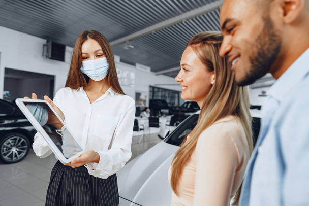 Car saleswoman wearing medical mask shows buyers couple something on digital tablet - Photo, Image