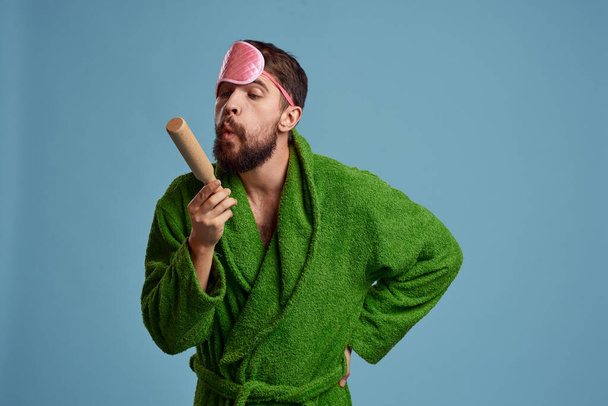 a man with a pink sleep mask holds a rolling pin in his hand and a green robe blue background emotions model - Photo, Image