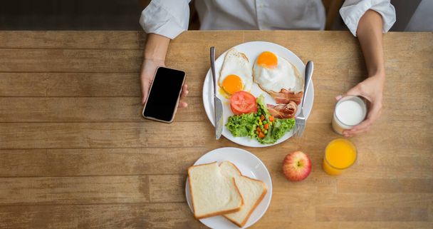 Здоровая женщина держит в руках смартфон и стакан свежего молока с завтраком на деревенском деревянном столе, наслаждаясь завтраком вместе, вид сверху, копия пробел-изображение - Фото, изображение