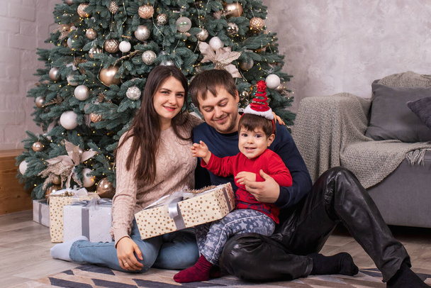 Mamá, papá e hijo pequeño se sientan cerca del árbol de Navidad y tienen regalos. Vacaciones familiares Año Nuevo, regalos y una mañana mágica - Foto, Imagen