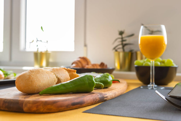 Snídaně v moderním, světlém interiéru - čerstvé bagety nakrájené na prkno, dvě papriky jalapeno, croissanty, hroznový a pomerančový džus ve sklenici - Fotografie, Obrázek