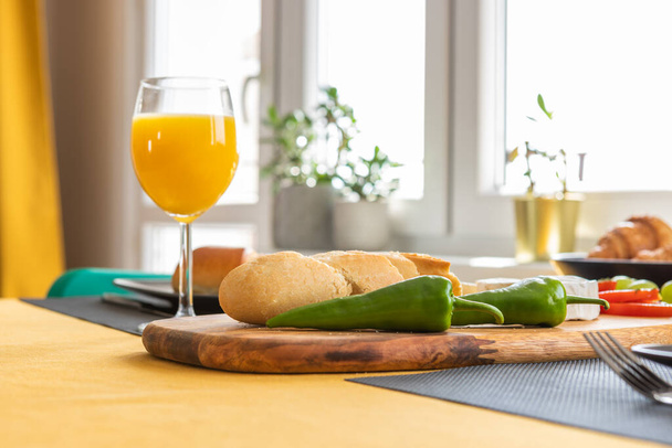 Breakfast in a modern bright interior - fresh baguette sliced on a board, two jalapeno peppers, croissants, mozzarella cheese and orange juice in glasses - Photo, Image