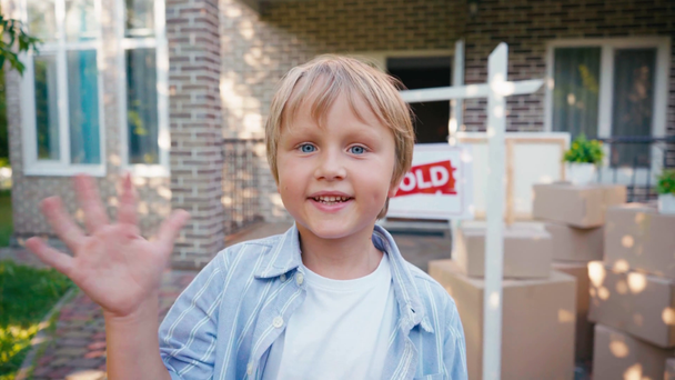 χαρούμενο αγόρι κουνώντας το χέρι και κοιτάζοντας την κάμερα κοντά στο νέο σπίτι - Πλάνα, βίντεο