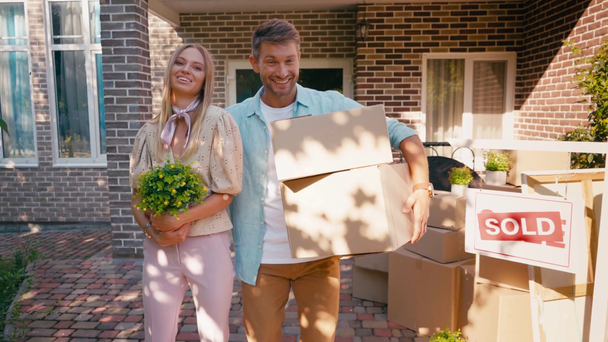 Zufriedenes Paar hält Kartons und Pflanze in der Nähe des neuen Hauses - Filmmaterial, Video