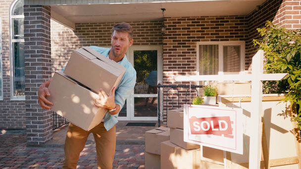 hombre apuntando con la mano al tablero vendido, sosteniendo cajas y saltando cerca de casa nueva - Imágenes, Vídeo