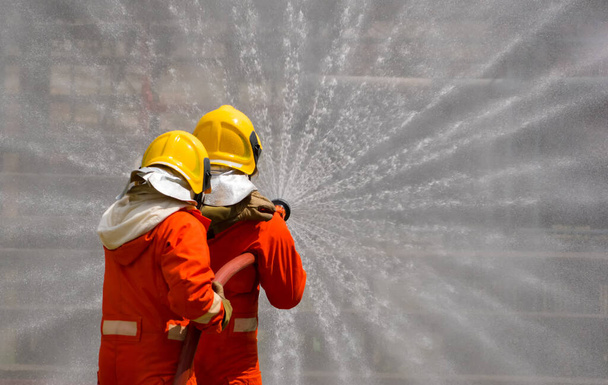 消火器と消火ホースからの水を使用して2人の勇敢な消防士、火災に高圧水を噴霧消防士、消防士の訓練、フォアグラウンドは水のスプリンガーのドロップです-画像 - 写真・画像