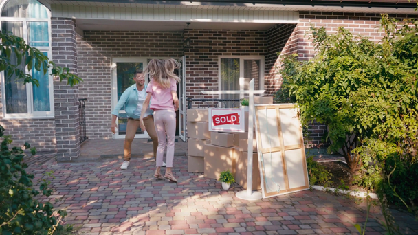 alegre pareja saltando y bailando cerca de cajas y nueva casa  - Imágenes, Vídeo
