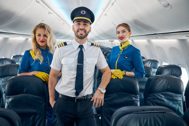 Glücklich lächelnde hübsche Pilotin mit zwei Stewardessen an Bord des Flugzeugs - Foto, Bild