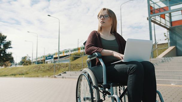 Joven mujer caucásica en silla de ruedas usando laptop. Tren pasando en el fondo - Foto, Imagen