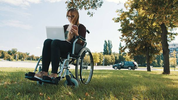 Távmunka koncepció. Fiatal fogyatékkal élő nő a kerekesszékben okostelefonnal és laptoppal - Fotó, kép