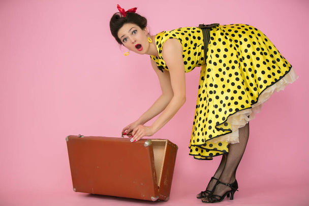 Beautiful pin-up girl opens a retro suitcase. Retro woman in yellow polka dot dress. The emotion of surprise. - Photo, Image