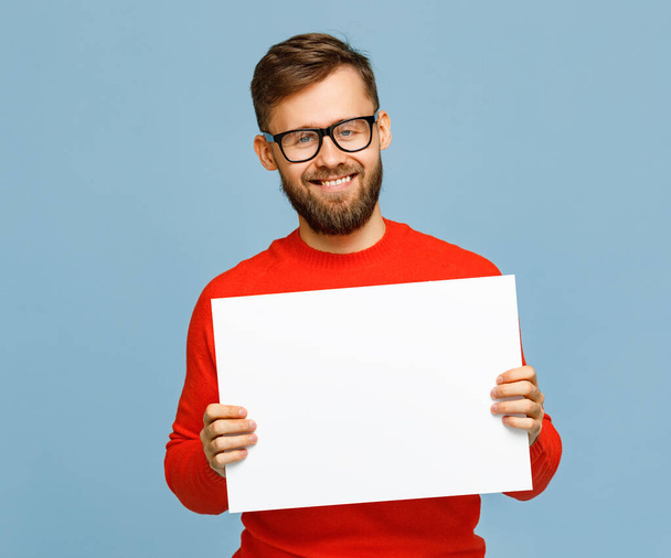 Positive bearded male in glasses smiling for camera and showing empty poster isolated on blue backgroun - Photo, Image