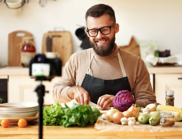 Happy γενειοφόρος άνθρωπος σε γυαλιά χαμογελώντας και να προετοιμάσει ένα χορτοφαγικό πιάτο ώριμα λαχανικά, ενώ γυρίσματα βίντεο για το μαγείρεμα vlog στην κουζίνα σε hom - Φωτογραφία, εικόνα