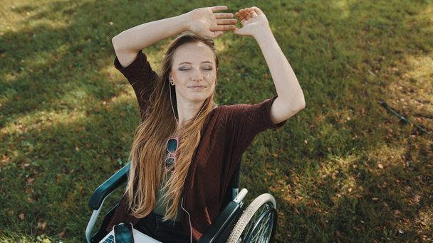 Молодая белая женщина в инвалидной коляске слушает музыку в наушниках в парке - Фото, изображение