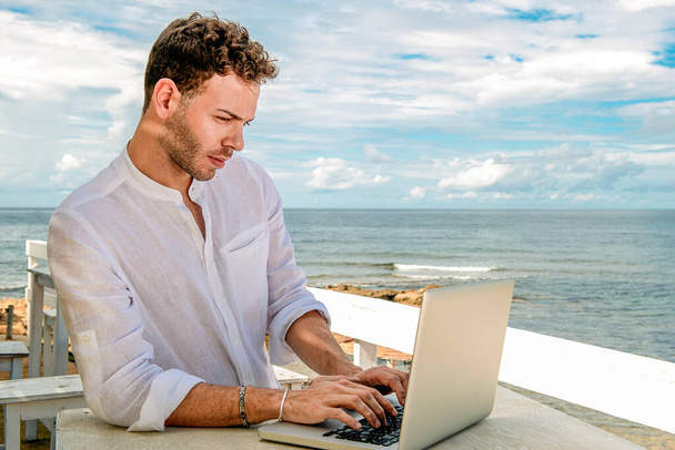 guapo y exitoso hombre caucásico trabajando con un portátil en la playa. trabajo independiente y remoto. estudiante en la orilla mediterránea - Foto, Imagen