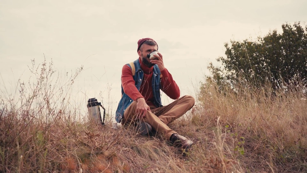 Traveler sitting and drinking tea from thermos on grass - Materiaali, video